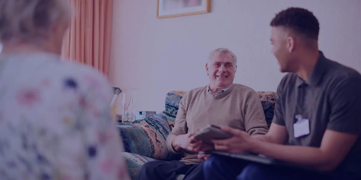 An older man and a younger man sit on a sofa, smiling and talking, with a woman in the foreground.