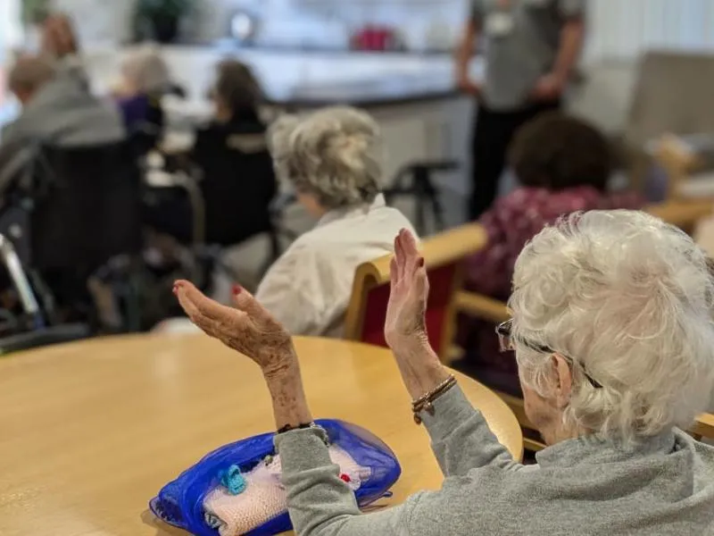 Elderly people, some in wheelchairs, sit in a care home, with one woman in the foreground clapping