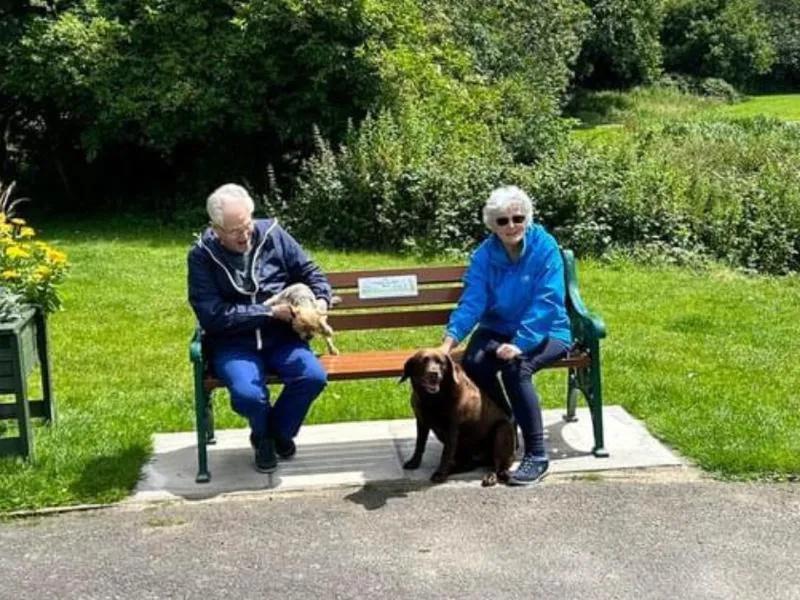 a man and his wife sat happily on one of Bury's Age Friendly benches in the park with their two dogs