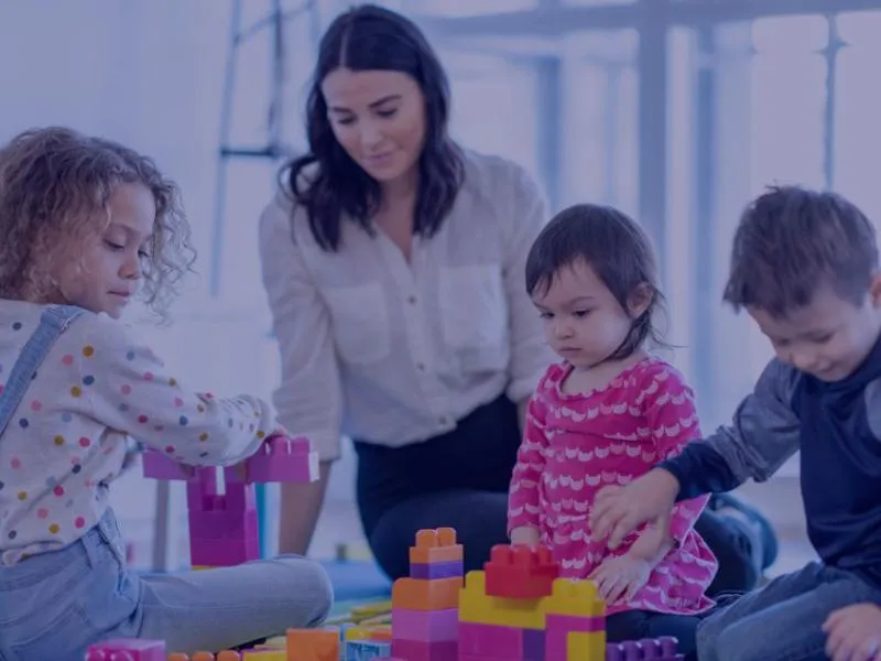 A group of young children and an adult playing together with large colorful building blocks in a bright, modern classroom.