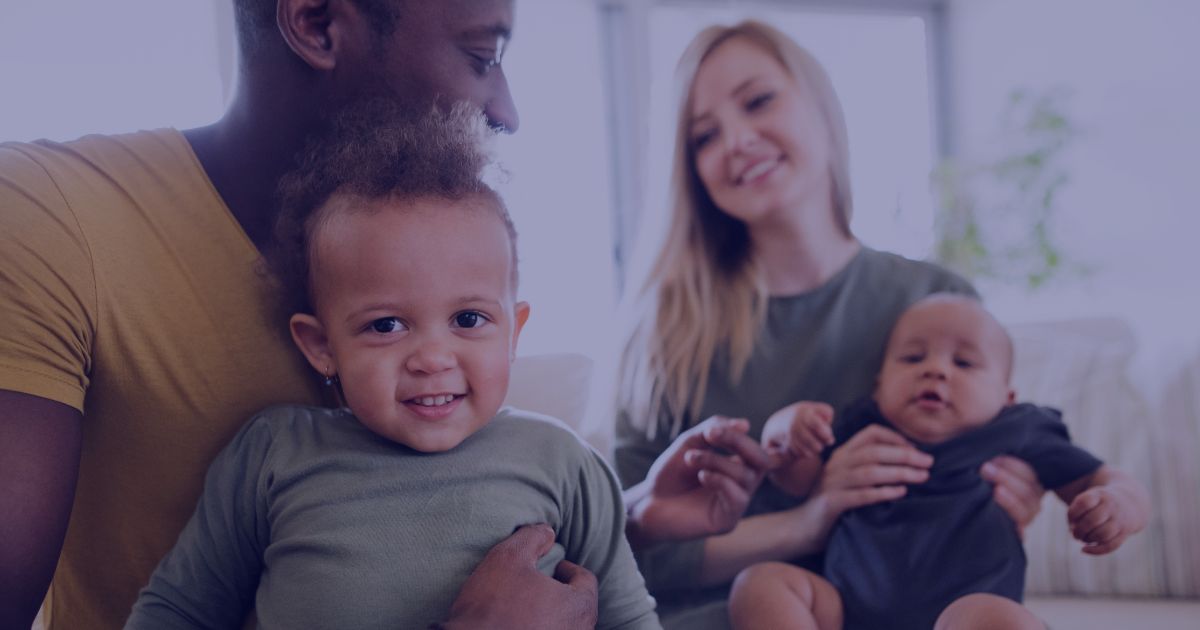 A smiling family sitting together indoors: a man holding a toddler and a woman holding a baby.