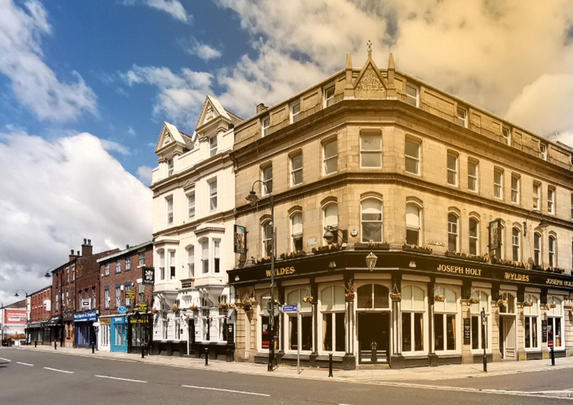 Street view of a traditional corner pub and neighbouring shops in a historic town centre, under a bright blue sky with scattered clouds