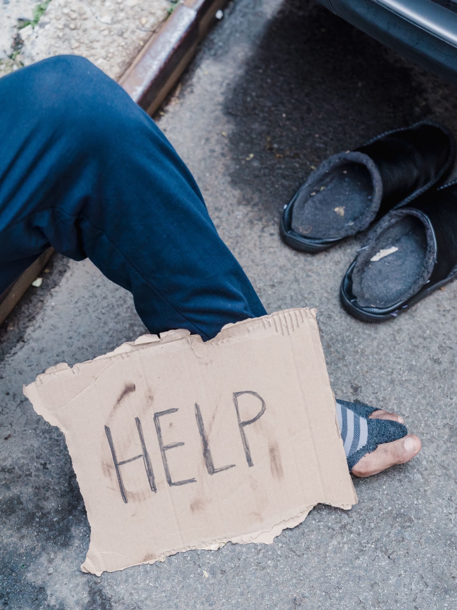 a handwritten sign with the word 'help' propped up by a mans leg wearing ripped clothing
