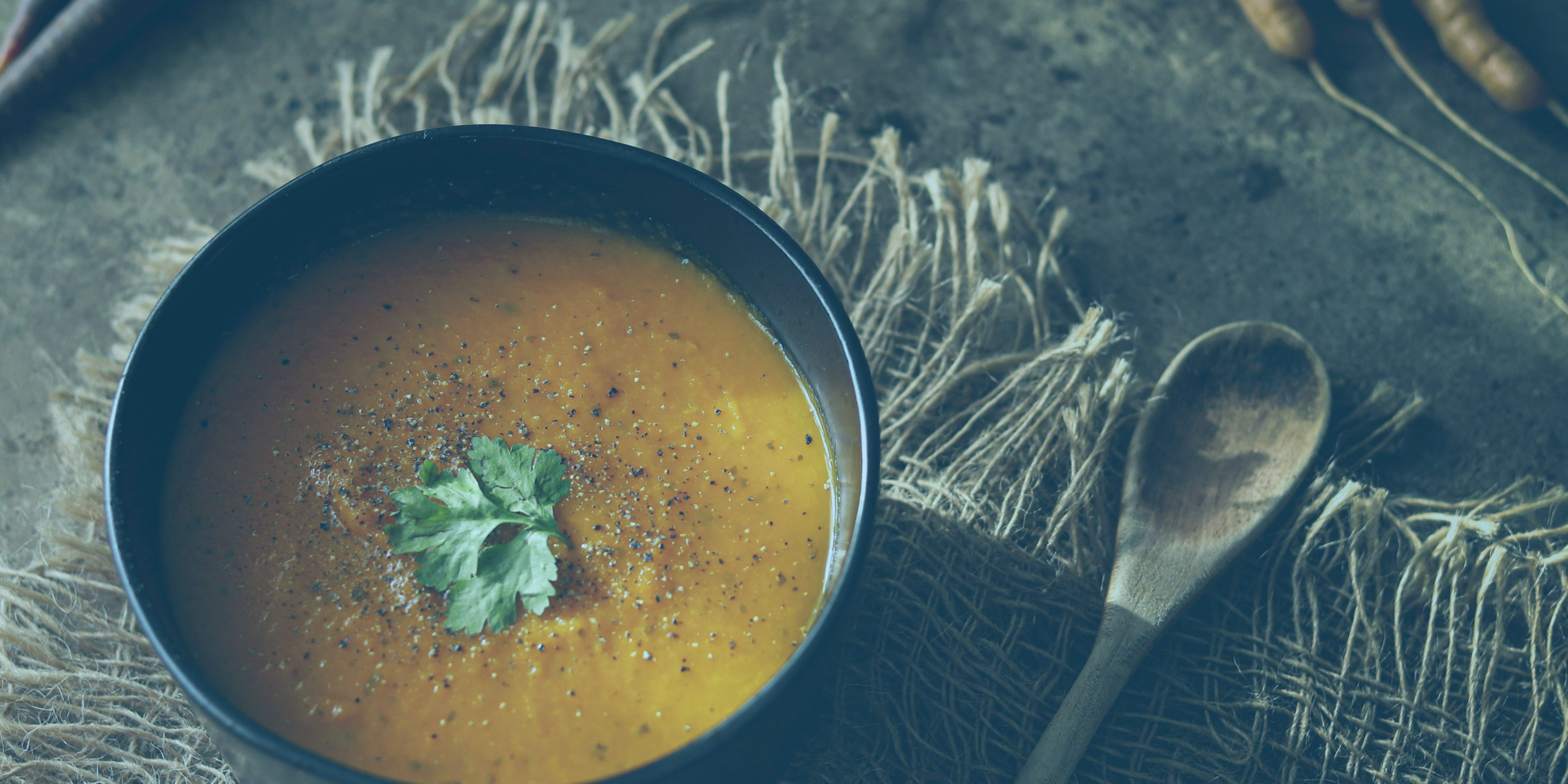 Bowl of orange soup garnished with fresh herbs, placed on a rustic surface with a wooden spoon beside it.