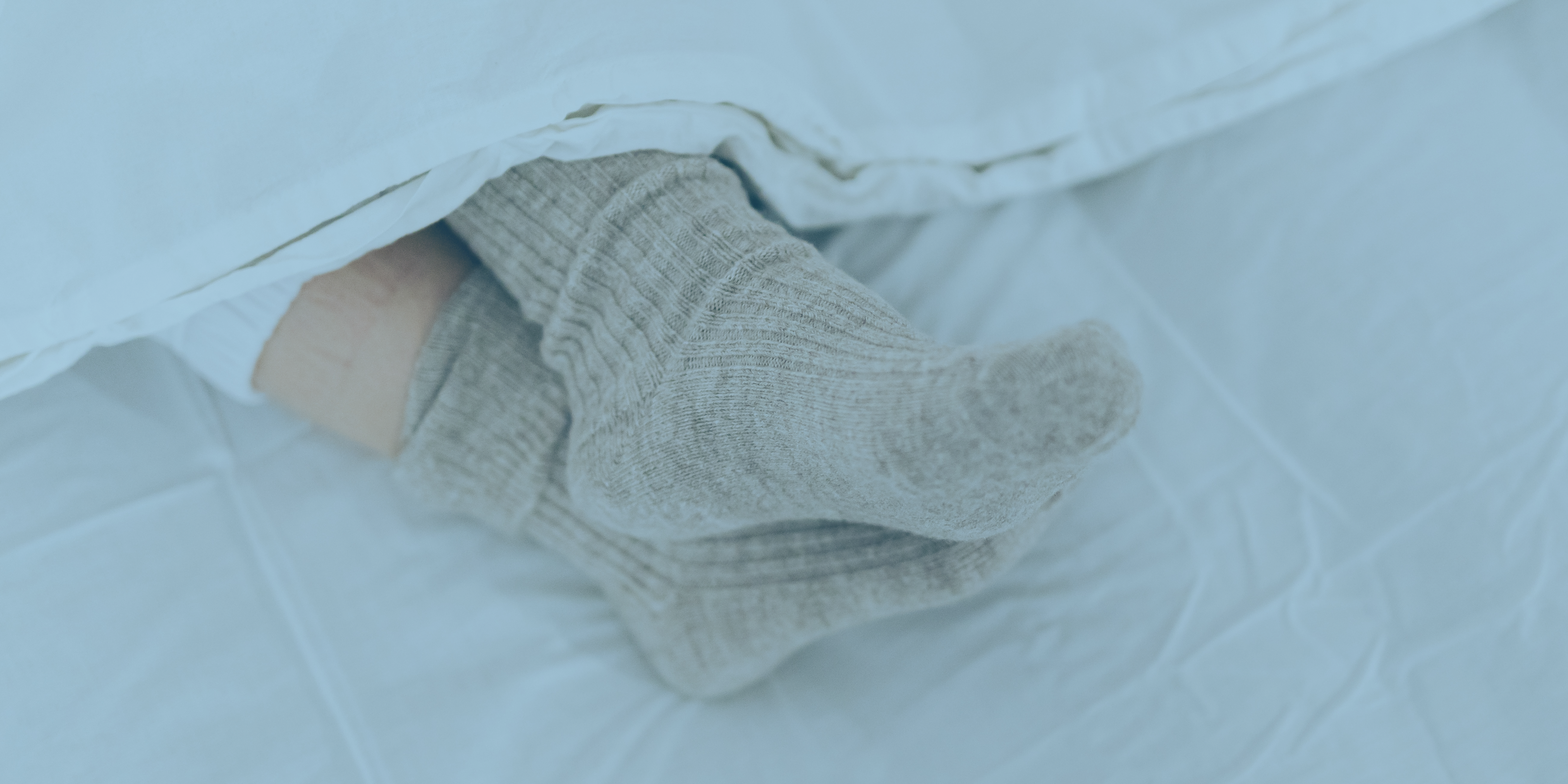 Close-up of feet wearing thick grey socks under a white blanket on a bed