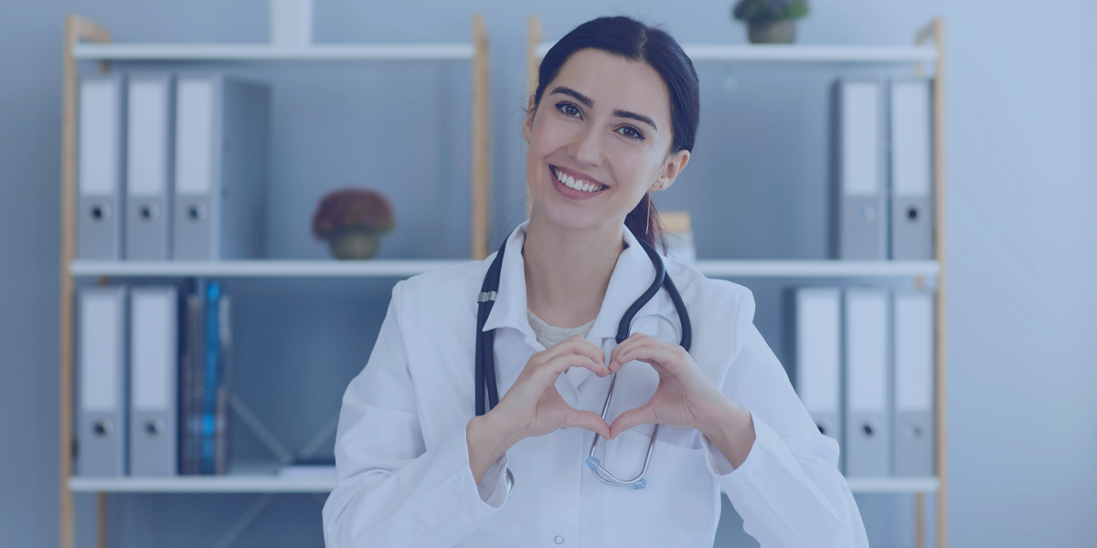 A woman in a doctor's coat with a stethoscope makes a heart shape with her hands and smiles.