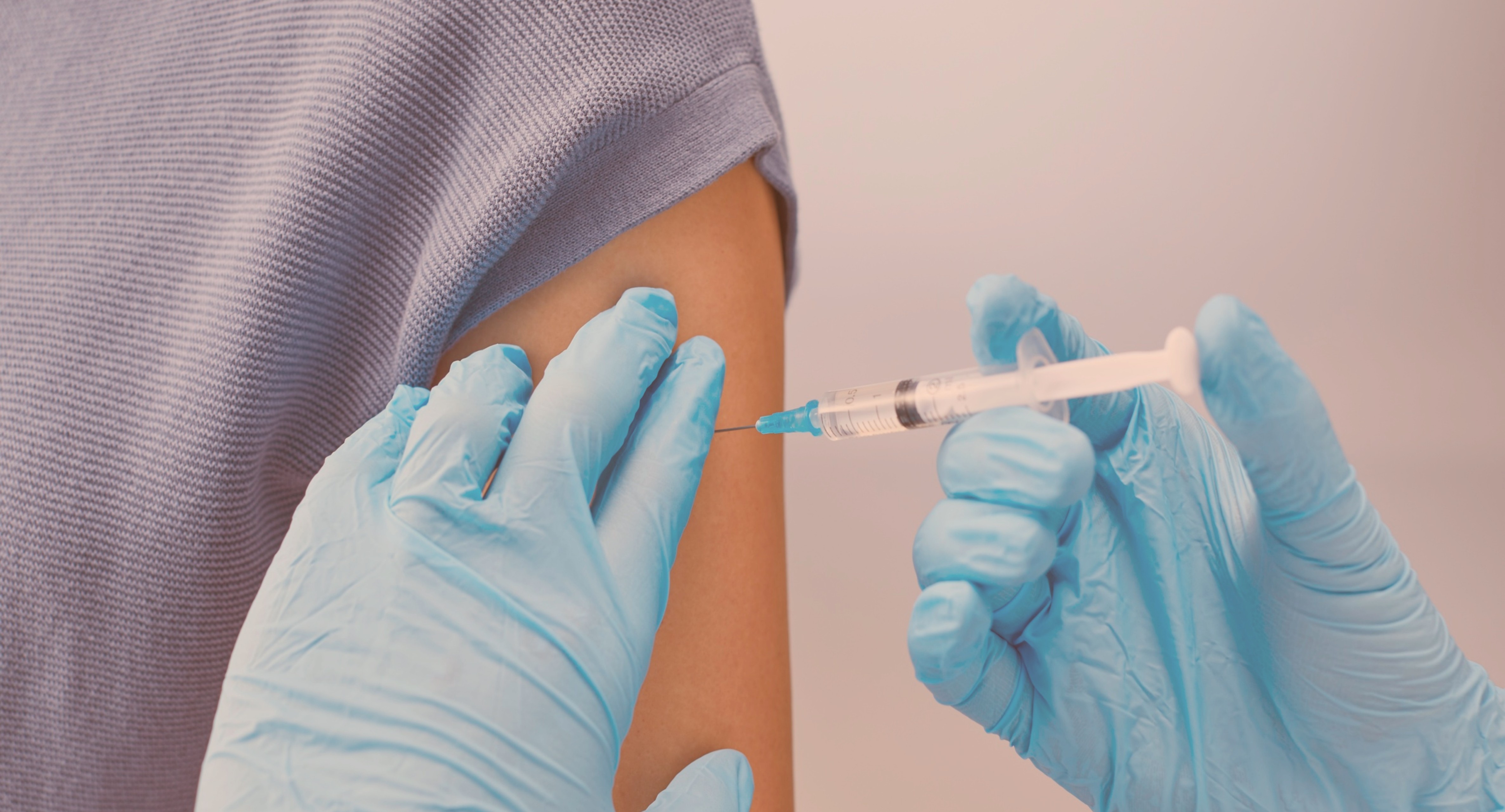 Healthcare professional wearing blue gloves administering a vaccination injection into a person’s upper arm.