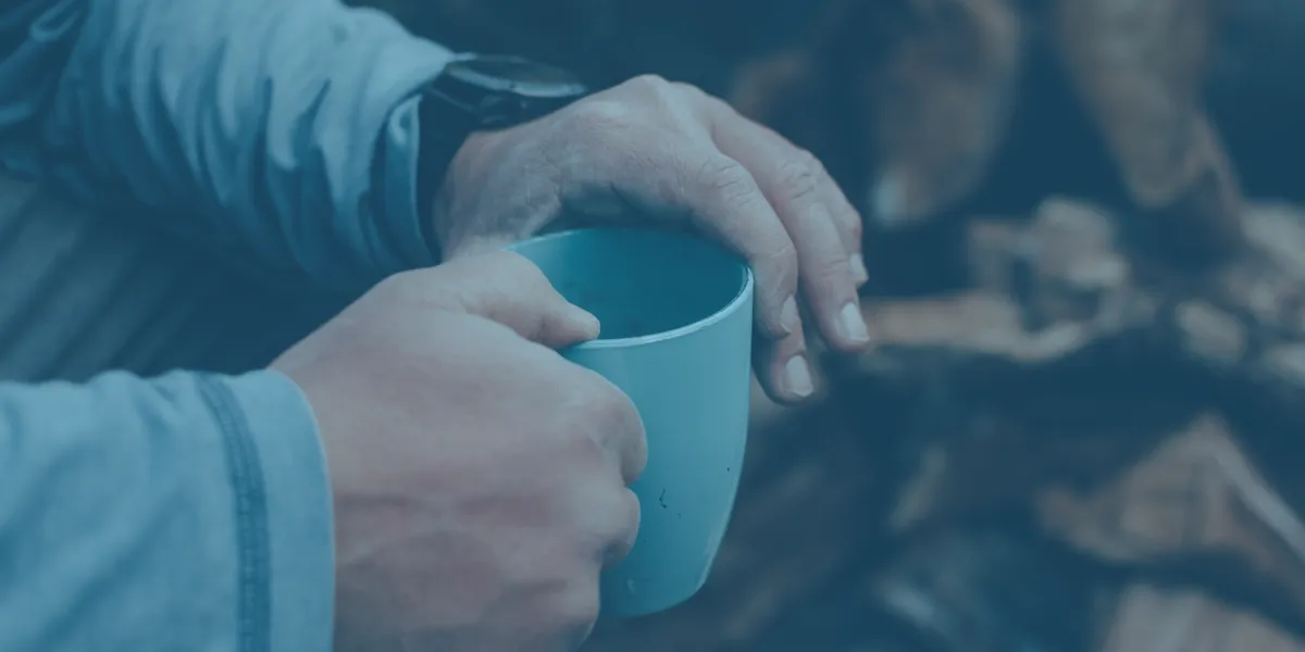 hands holding a light blue mug, with a wristwatch visible and chopped wood in the background