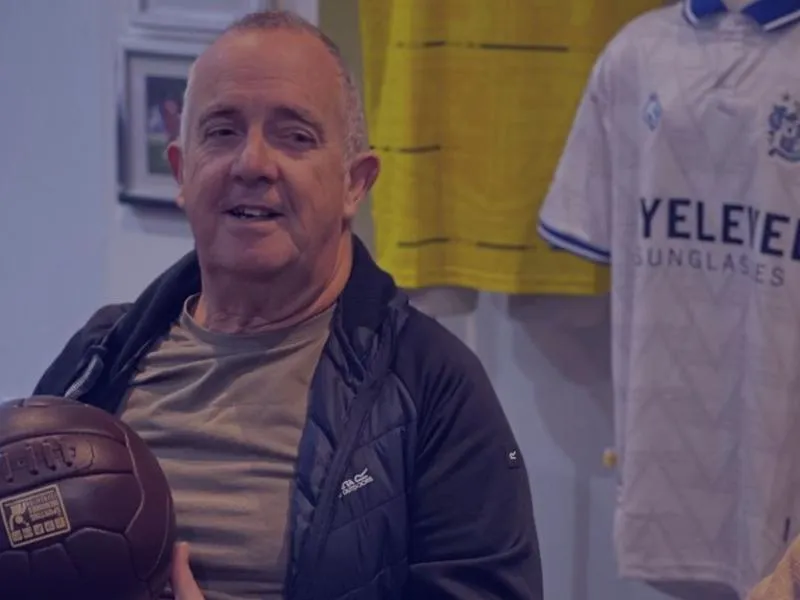 a man in a group setting smiling and holding an old football whilst surrounded by sporting memorabilia
